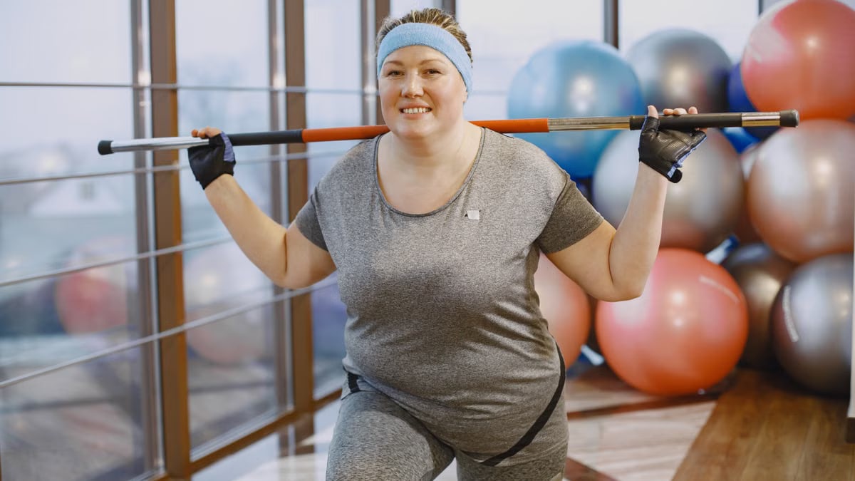 A plus-size woman in gym attire exercising with a barbell in a modern gym environment.